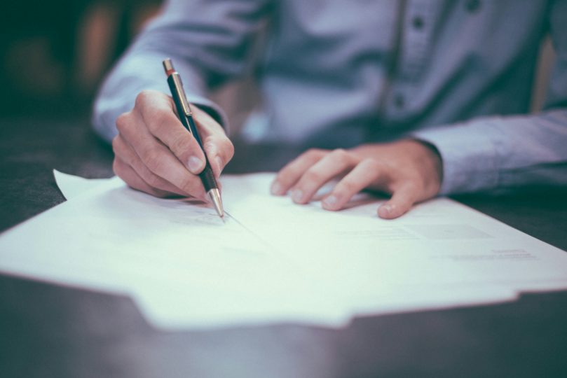 A person in a blue shirt writing on documents at a brown table. 